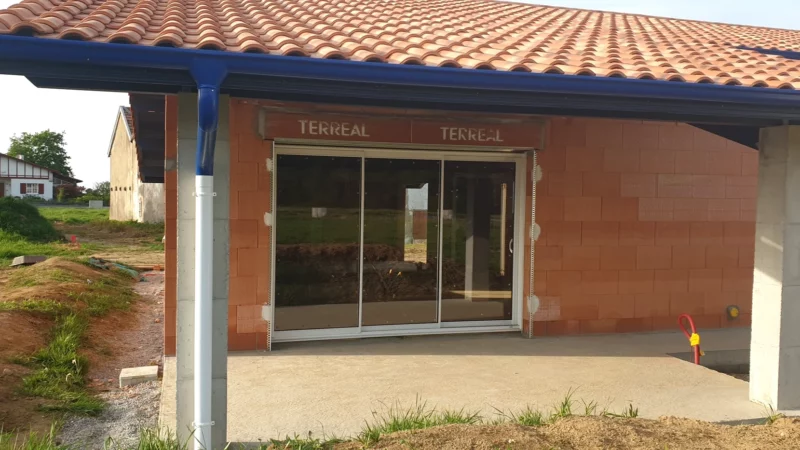 Baie vitrée posée sous la terrasse couverte d’une maison neuve en briques.