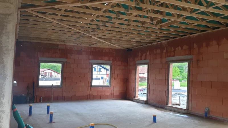 Intérieur d’une maison neuve montrant la charpente bois et l’écran sous-toiture au-dessus des murs en briques.
