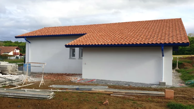 Façade latérale enduite d’une maison neuve avec toiture en tuiles et gouttières bleues.