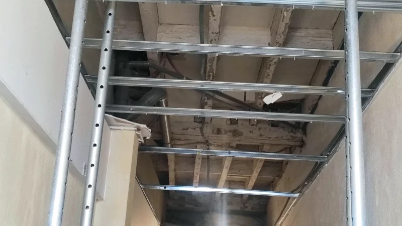 Solives anciennes visibles derrière une ossature métallique de plafond étayée.